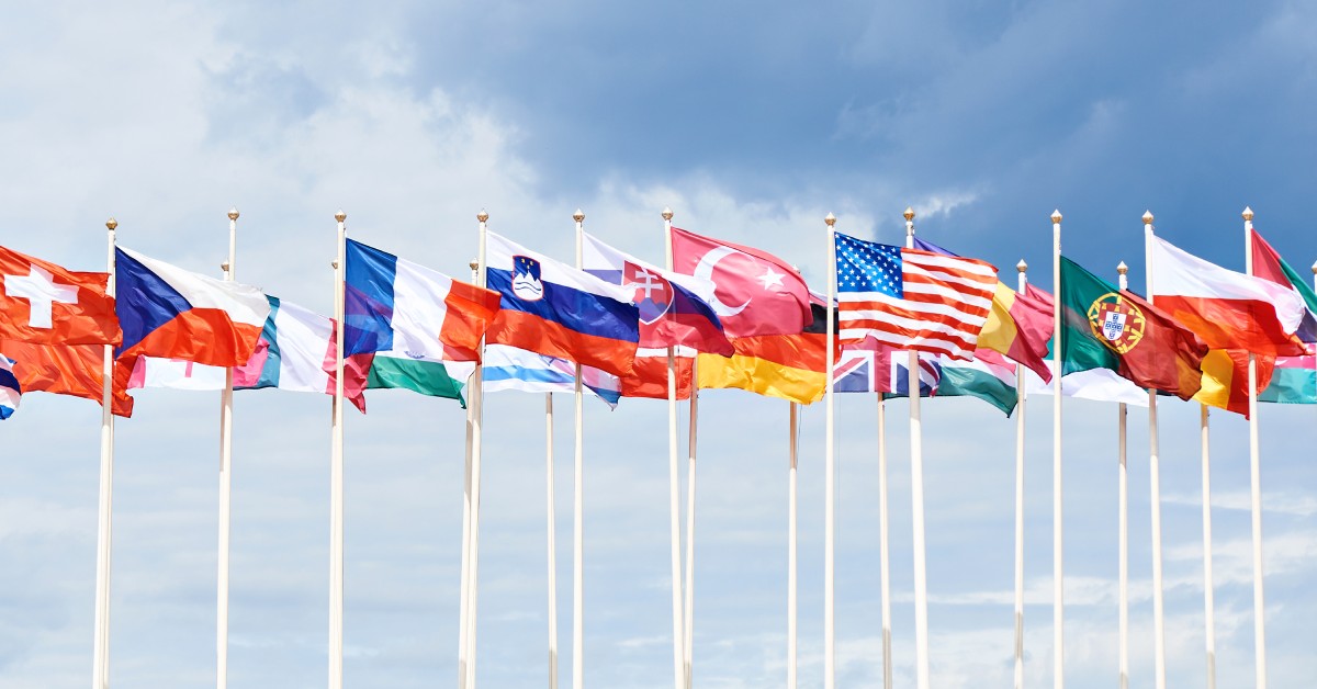 Various country flags on poles against a cloudy sky background including flags of the US, Sweden, Japan, Turkey, and more.