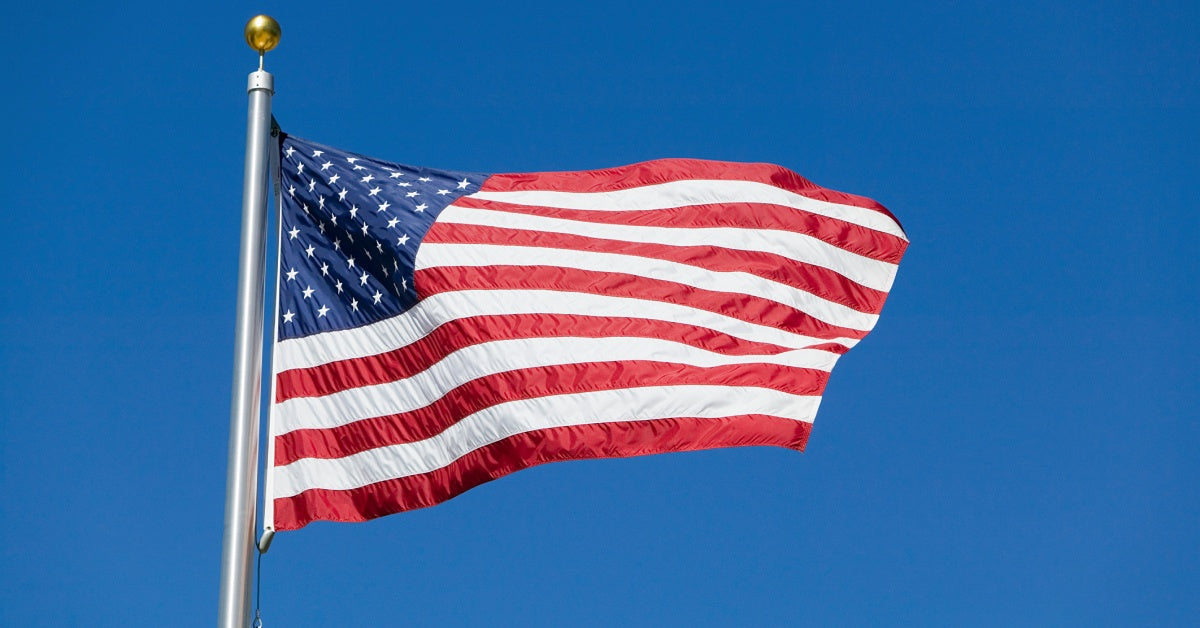 A red, white, and blue American flag flying on a large, silver flagpole with a clear blue sky behind it.