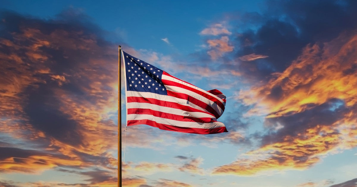 A US flag is flying on a pole with a colorful blue sky and orange, red, and dark clouds behind it.