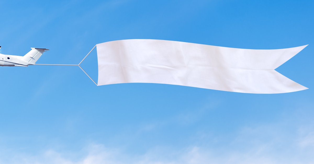 A small white airplane towing a large, blank white advertising banner through a blue sky via a short rope.