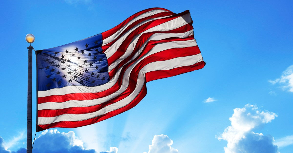 A large American flag flying on a large flagpole. Behind, the sun shines through the fabric with clouds in the sky.