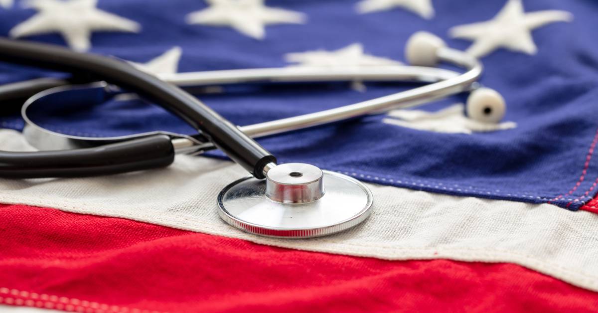 A close-up of a stitched American flag with a medical stethoscope resting on red and white stripes and blue and white stars.