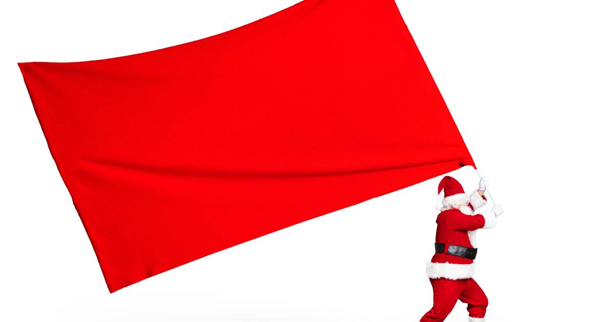 A small Santa Claus holds a large, blank red and white flag that's twice his size against a white background.