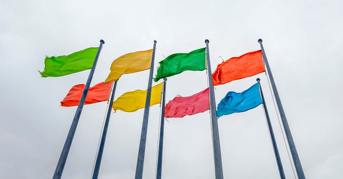 Eight flags in different colors, including orange, yellow, red, green, and blue, on flagpoles flap in the breeze.