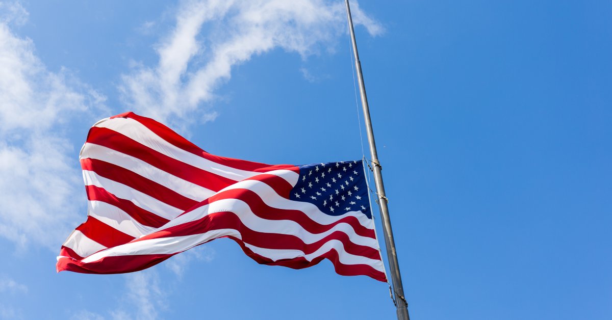 An American flag hung at half-staff from a silvery flagpole against a bright blue sky with a few wispy clouds.