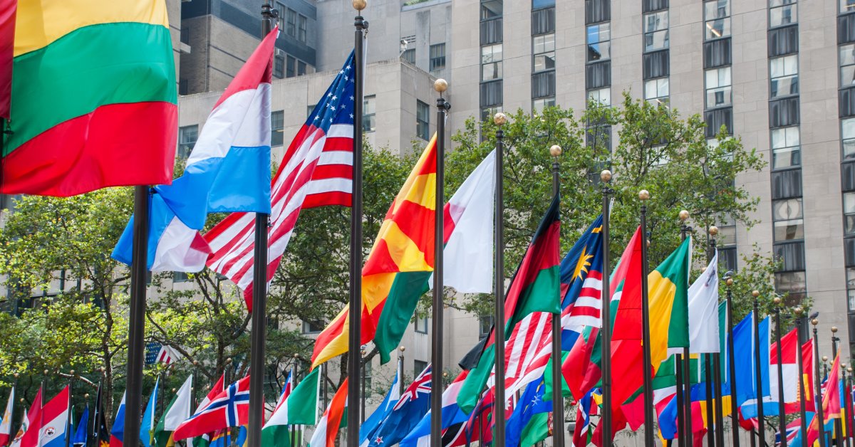 A circle of flags of all nations atop different flagpoles, flapping outside with large buildings behind them.