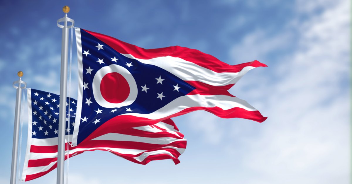 The Ohio state flag and American flag are seen flapping against a bright blue sky with a few wispy clouds.