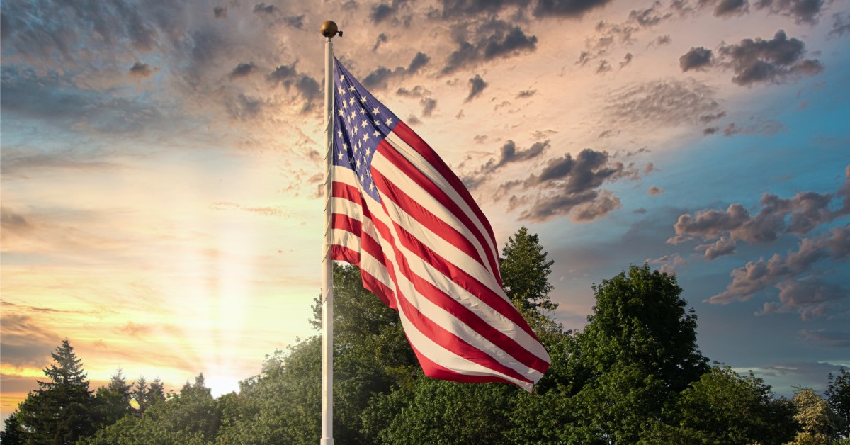 A tall white flagpole holds a slightly furled American flag. The sun sets in the background against a dusky sky.